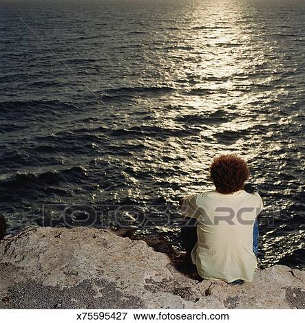 Man Sitting On Edge Of Cliff Looking At Ocean Rear View Man Sitting On Edge Of Cliff Looking At Ocean Rear View