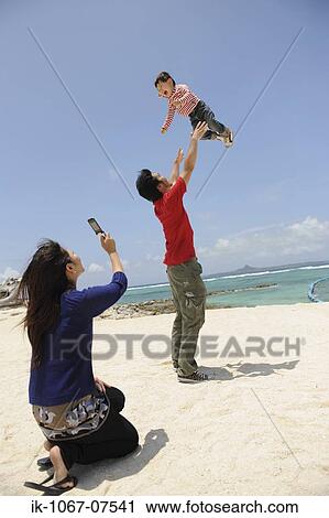 Stock Image - Mother taking snaps while father lifting child into air. Fotosearch