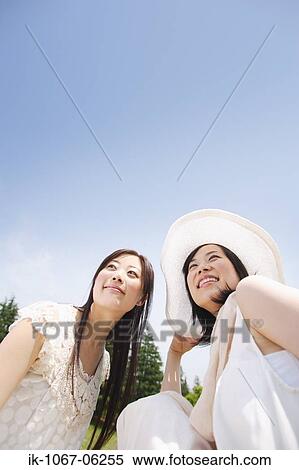 Stock Photography - Two young smiling women sitting against blue sky. Fotosearch