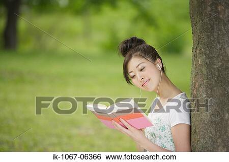 Stock Photograph - Young woman listening music leaning against tree. Fotosearch