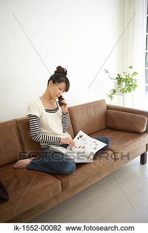 Stock Image - Young woman reading magazine and talking on cell phone. Fotosearch