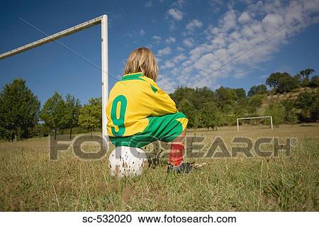 Stock Image - Boy in soccer uniform sitting on ball near goalpost . Fotosearch