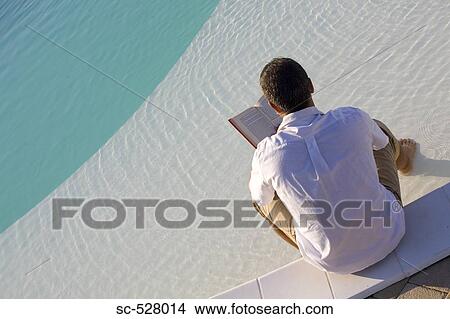 Picture - Man sitting at edge of pool and reading book. Fotosearch
