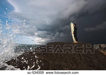 Stock Photo - Man standing on rock with his surfboard against cloudy sky. Fotosearch