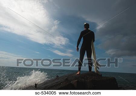 Stock Photography - Man standing on rock with surfboard looking at ocean . Fotosearch
