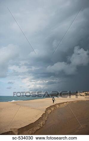 Stock Image - Man walking on sand with his surfboard on stormy day. Fotosearch