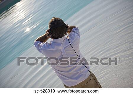 Stock Photo - Man wearing headphones and listening to music by poolside. Fotosearch