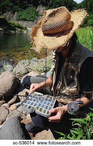 Picture - Man with straw hat sitting on stone and holding fly box. Fotosearch