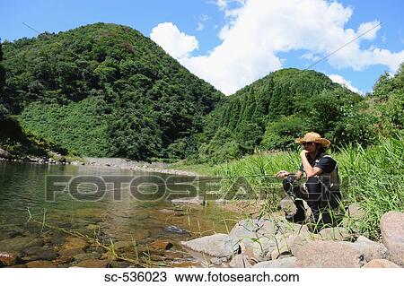 Stock Image - Man with straw hat sitting on stone and holding fishing rod. Fotosearch
