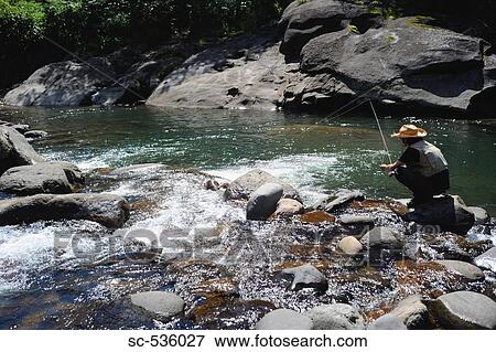 Stock Photo - Man with straw hat sitting on stone and fishing. Fotosearch