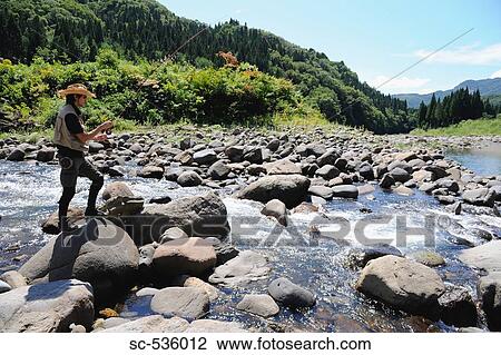 Stock Image - Man with straw hat standing on stone and holding fishing rod. Fotosearch