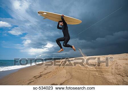 Stock Image - Surfer carrying surfboard on head and jumping up in air. Fotosearch