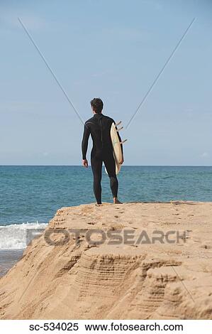 Stock Photography - Surfer standing on rock and holding surfboard under his arm. Fotosearch