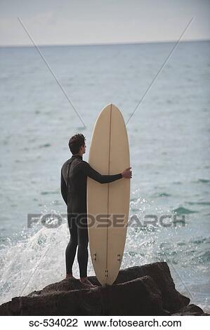 Stock Image - Surfer standing on rock with surfboard looking at sea . Fotosearch