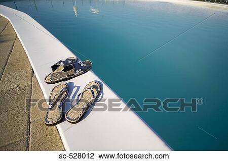 Stock Image - Two pair of flip flops at the edge of swimming pool. Fotosearch