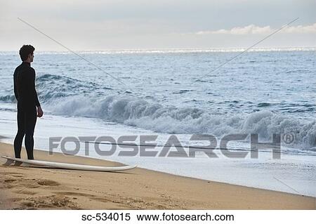 Young adult in wetsuit standing on beach along shoreline View Large Photo Image Stock Photography - Young adult in wetsuit standing on beach along shoreline. Fotosearch
