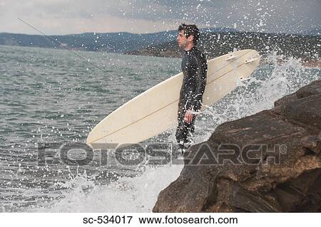 Stock Photo - Young adult standing near rock holding his surfboard. Fotosearch