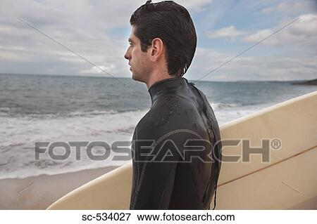 Stock Photo - Young man in wetsuit holding his surfboard. Fotosearch