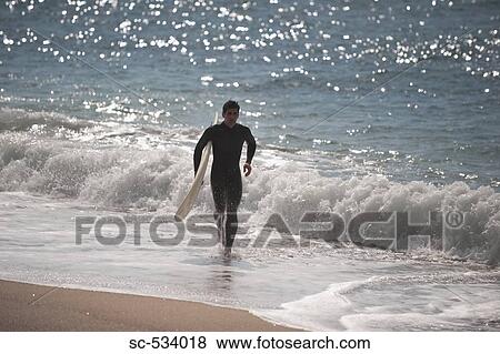 Stock Photo - Young man running along shoreline with his surfboard. Fotosearch