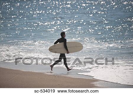 Stock Photo - Young man running toward sea with his surfboard. Fotosearch