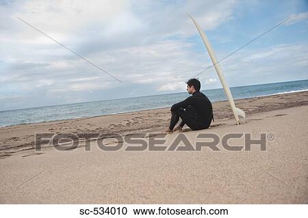 Stock Image - Young man sitting on beach with his surfboard. Fotosearch