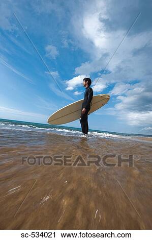 Stock Image - Young man standing with his surfboard near shoreline. Fotosearch