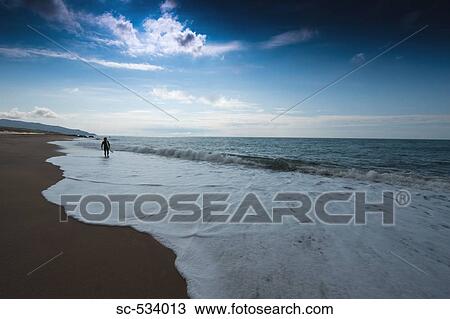 Stock Image - Young man walking along shoreline with his surfboard. Fotosearch