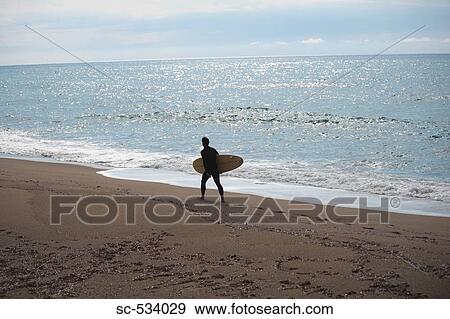 Stock Photo - Young man walking along shoreline with his surfboard. Fotosearch