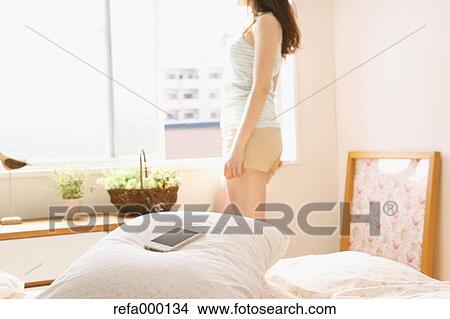 Picture - Smart phone on pillow and young woman by the window in the background. Fotosearch