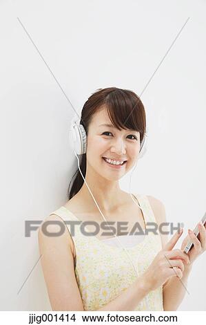 Picture - Japanese young woman in a one piece dress with earphones standing against white background. Fotosearch