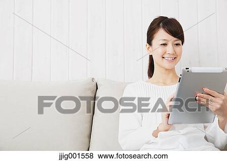 Stock Photo - Japanese young woman in a white shirt with tablet on the sofa. Fotosearch