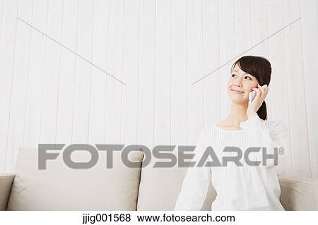 Stock Photo - Japanese young woman in a white shirt with smartphone on the sofa. Fotosearch