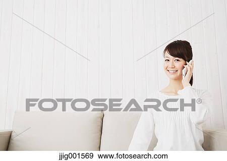 Stock Photo - Japanese young woman in a white shirt with smartphone on the sofa. Fotosearch