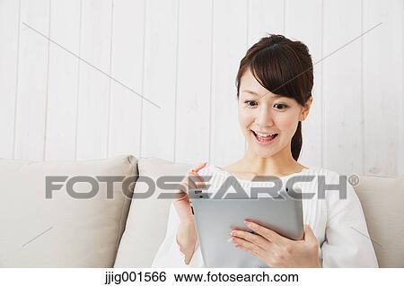 Stock Photograph - Japanese young woman in a white shirt with tablet on the sofa. Fotosearch