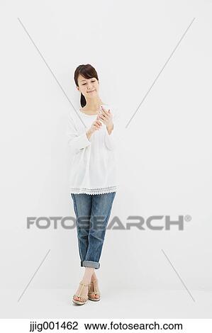 Stock Image - Japanese young woman in jeans and white shirt with smartphone standing against white background. Fotosearch