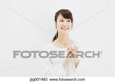 Stock Image - Japanese young woman in jeans and white shirt with earphones standing against white background. Fotosearch