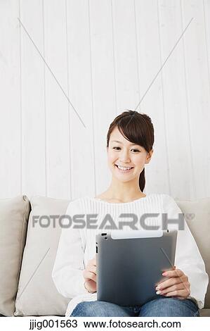 Stock Image - Japanese young woman in jeans and white shirt with tablet on the sofa. Fotosearch