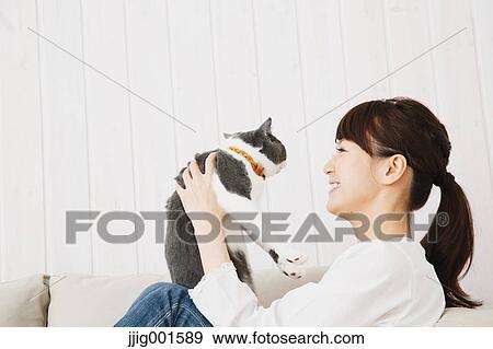 Stock Photo - Japanese young woman in jeans and white shirt with cat on the sofa. Fotosearch