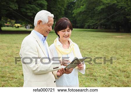 Stock Image - Senior adult Japanese couple with tablet in a park. Fotosearch