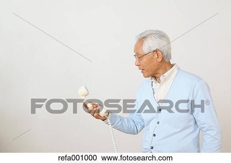 Stock Image - Senior adult Japanese man with phone against white wall. Fotosearch