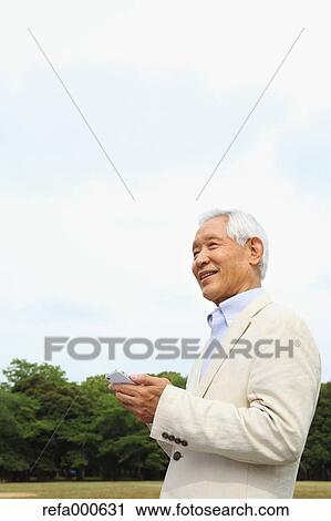 Stock Image - Senior adult Japanese man with smartphone in a park. Fotosearch