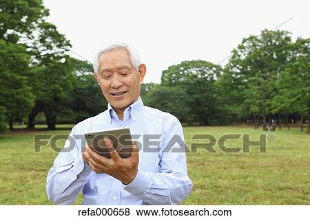 Stock Photo - Senior adult Japanese man with tablet in a park. Fotosearch