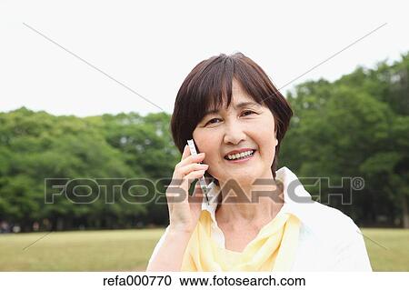 Stock Image - Senior adult Japanese woman with smartphone in a park. Fotosearch