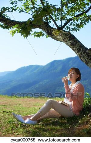 Young Japanese woman sitting with book in the countryside drinking water View Large Photo Image Stock Photo - Young Japanese woman sitting with book in the countryside drinking water. Fotosearch