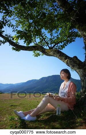 Stock Photography - Young Japanese woman sitting with book in the countryside. Fotosearch