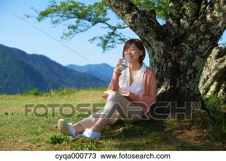 Stock Image - Young Japanese woman sitting with water and book in the countryside. Fotosearch