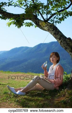 Stock Photo - Young Japanese woman sitting with water and book in the countryside. Fotosearch