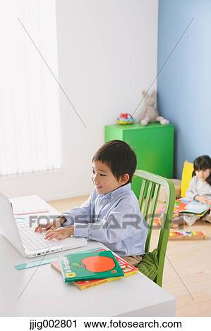 Stock Image - Elementary age boy doing his homework at his desk with his sister reading on the floor in the background. Fotosearch