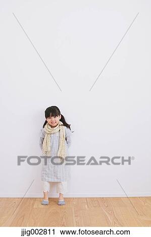 Stock Image - Elementary age girl leaning against a white wall and smiling at camera. Fotosearch