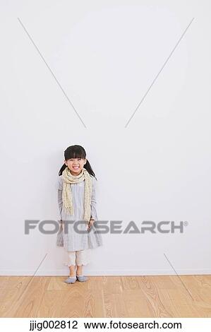 Stock Image - Elementary age girl leaning against a white wall and smiling at camera. Fotosearch